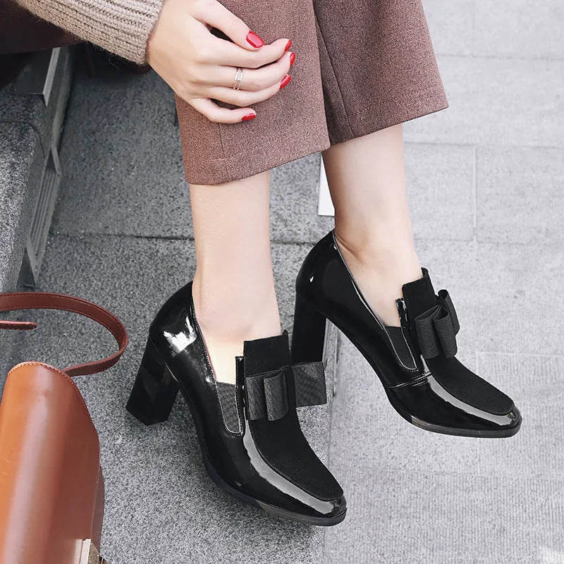 Black patent leather women's high heels with a thick super high heel and square toe on a plain background.