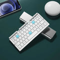 Silver white mini folding Bluetooth keyboard open next to a smartphone on its integrated stand.