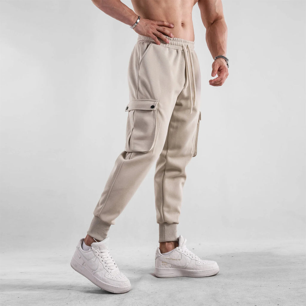 Men's loose fit beige cotton trousers with an ankle tie and multiple pockets on a man standing casually.