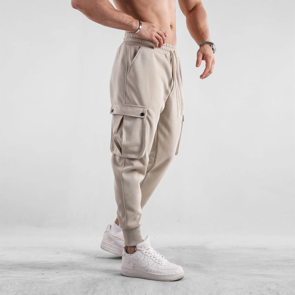 Men's loose fit beige cotton trousers with an ankle tie and multiple pockets on a man standing casually.