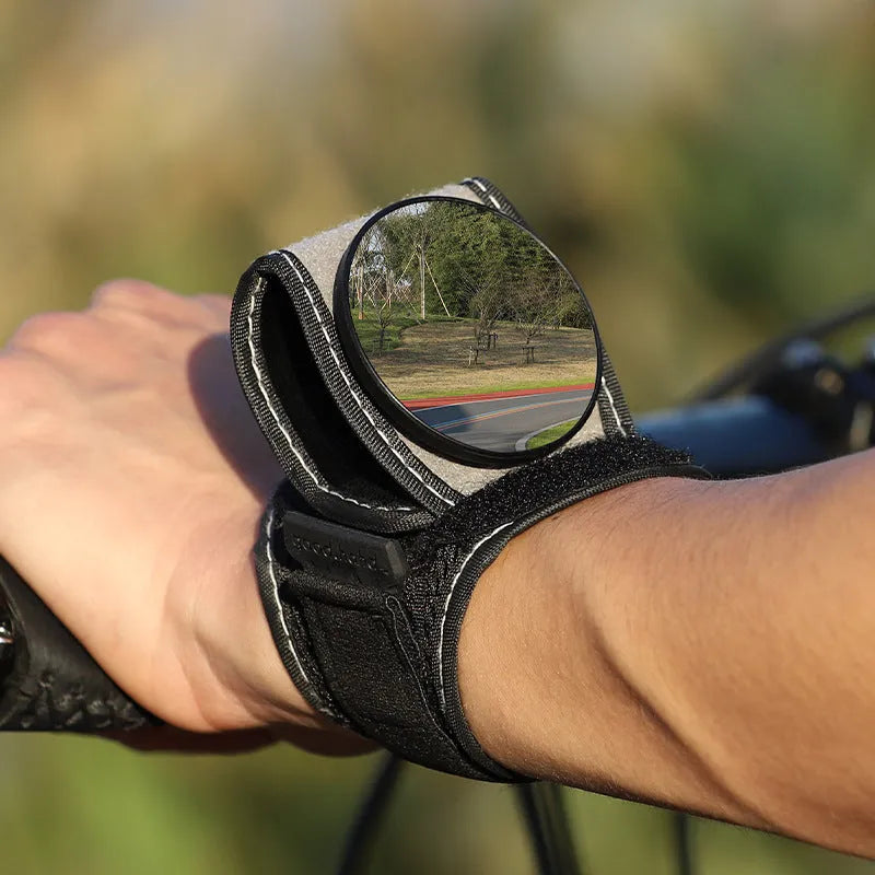 Black nylon wrist-mounted bicycle rear-view mirror shown on a cyclist's wrist.