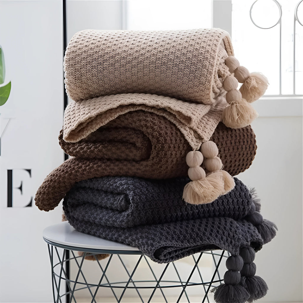 Nordic style acrylic knit blanket with fringed ball trim in a choice of four colours on a sofa.