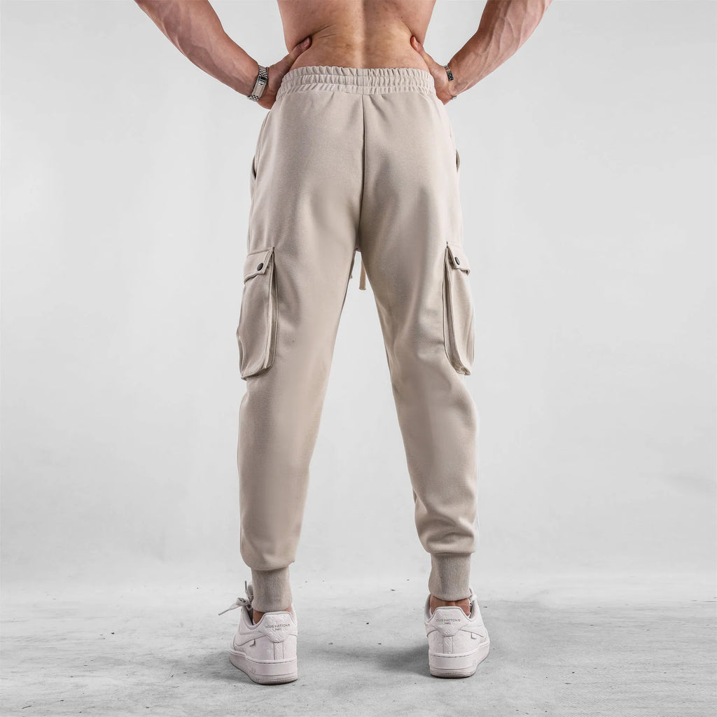 Men's loose fit beige cotton trousers with an ankle tie and multiple pockets on a man standing casually.